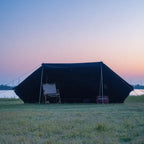 Large black camping tent with a folding chair on grassy field by a river at sunset